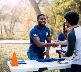 A man with a blue shirt shaking hands and gifting another man a blue shift outside in the park while a woman watches.