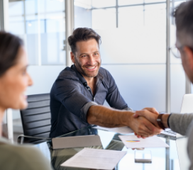 A man shaking hands with another man across a conference table in an office with a glass table and documents on the desk.