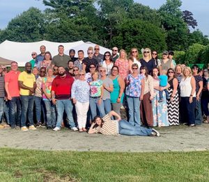 A large group of employees posing for a photo outside in front of a big white tent, some wearing sunglasses and others in tie dye or summer prints.