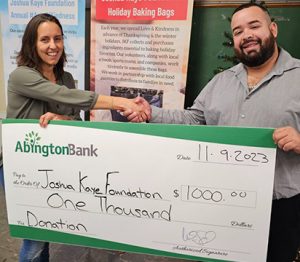 A man and woman shaking hands while holding a large check made out to the Joshua Kaye Foundation.