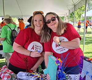 Two woman in red shirts leaning towards eachother behind a table under a tent that has a variety of oven mitts on it and people all around them.